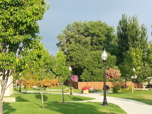 A serene park scene with green trees, colorful flowers, and lampposts lining a winding path under a cloudy sky.