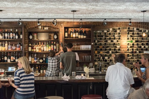 A cozy bar scene with patrons enjoying drinks, a bartender serving, and shelves lined with various bottles.