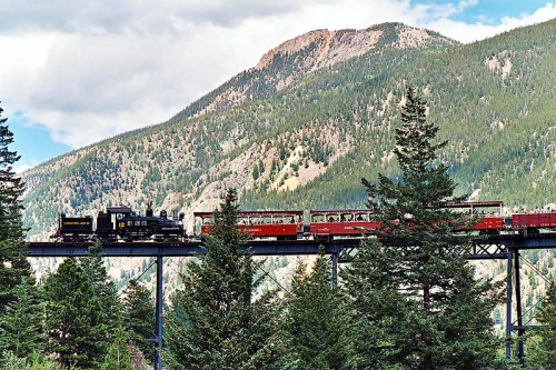 A vintage train crosses a bridge surrounded by lush trees and mountains under a partly cloudy sky.