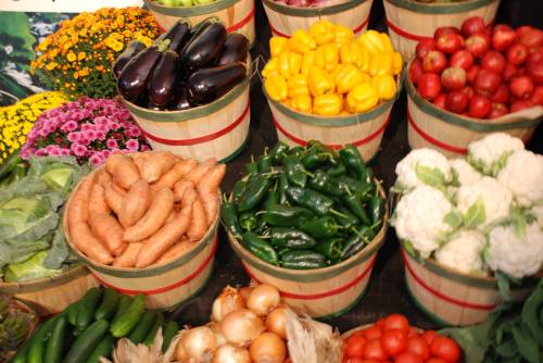 A vibrant display of fresh vegetables in baskets, including sweet potatoes, peppers, cauliflower, and various herbs.