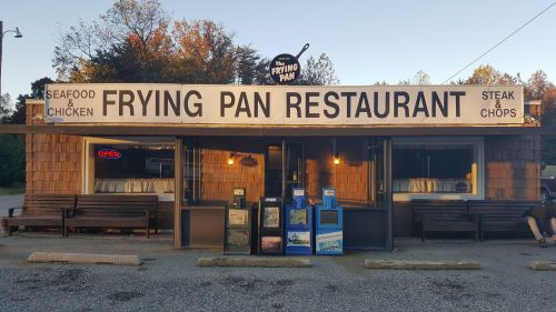 Exterior of the Frying Pan Restaurant, featuring a sign with "Seafood & Chicken" and "Steak & Chops," with benches outside.