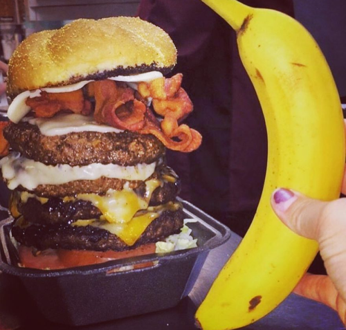 A towering burger with multiple patties, cheese, and bacon, next to a hand holding a large banana.