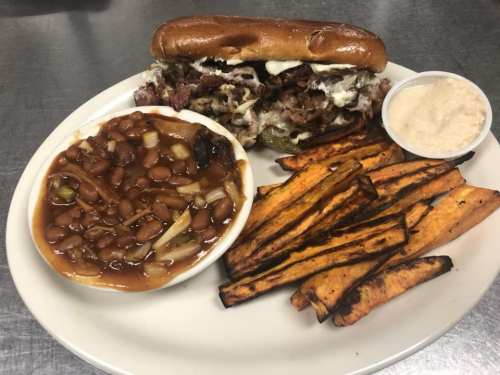 A plate featuring a loaded sandwich, sweet potato fries, baked beans, and a small cup of coleslaw.