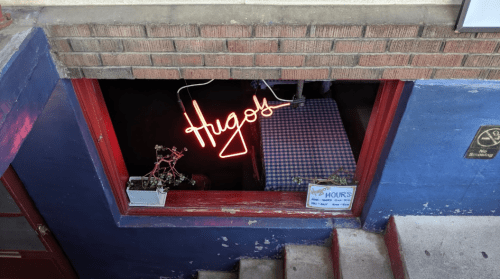 A neon sign reading "Hugo's" in a window, with a checkered tablecloth and small plants visible inside.