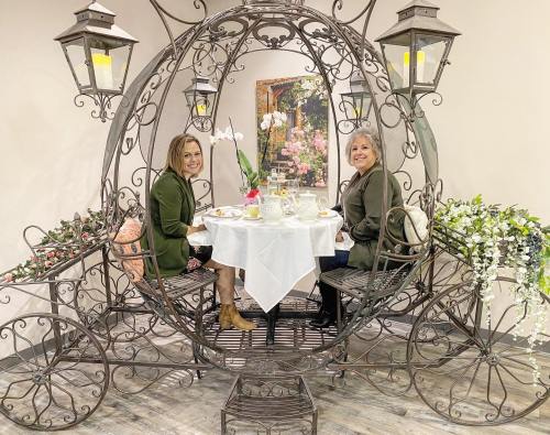 Two women sit at a table in a decorative carriage, surrounded by flowers and lanterns, enjoying tea together.
