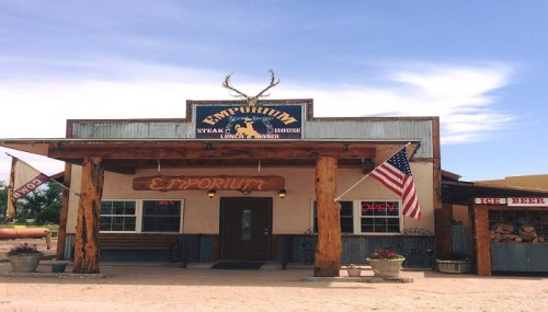 A rustic steakhouse with a wooden facade, American flag, and antlers above the entrance, set against a blue sky.