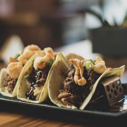 Three tacos filled with shredded meat, topped with crispy onions and green onions, served on a black plate.