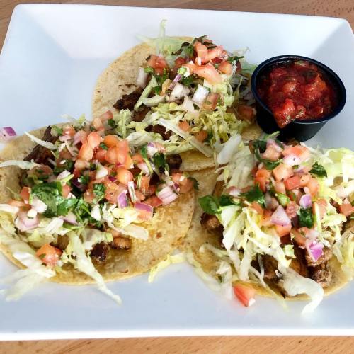 Three tacos topped with lettuce, diced tomatoes, and onions, served with a side of salsa on a white plate.