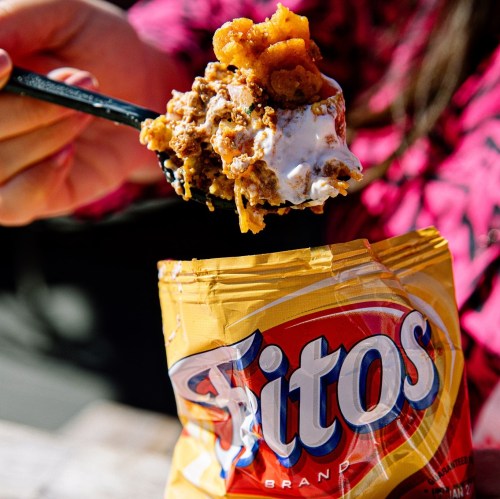 A hand holding a spoonful of a layered dish with Fritos, topped with a creamy mixture, next to an open Fritos bag.
