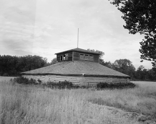 A round, weathered wooden building with a flat roof, surrounded by tall grass and trees under a cloudy sky.