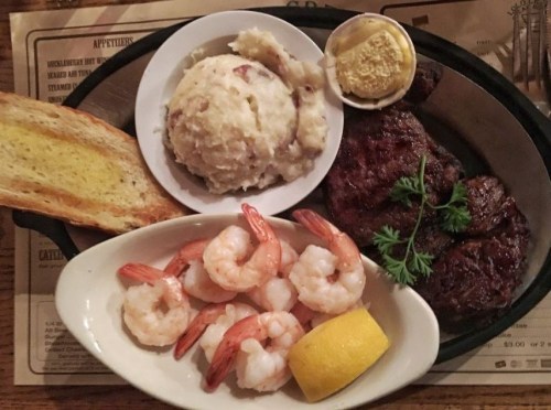 A plate featuring grilled steak, shrimp, mashed potatoes, toast, and a lemon wedge, garnished with parsley.