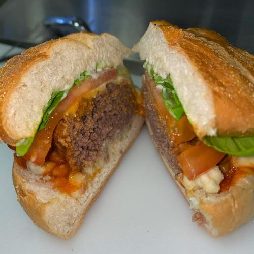 A close-up of a sliced sandwich with a beef patty, lettuce, tomato, and sauce on a toasted bun.