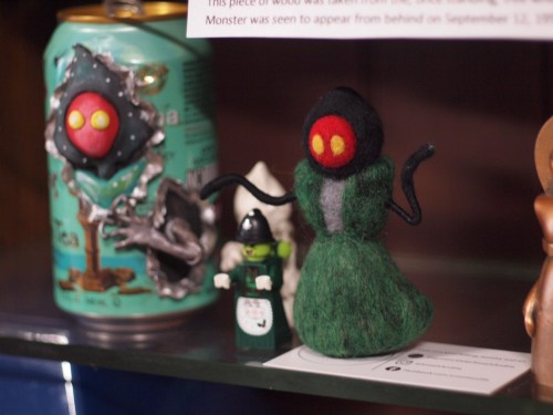 A green felted figure with red eyes and black accents stands beside a can and a small Lego figure on a shelf.