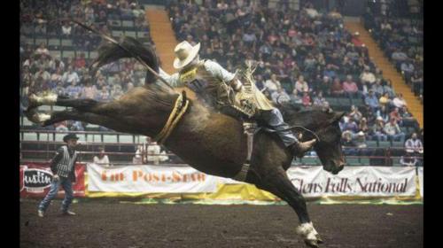 A cowboy rides a bucking horse in a rodeo arena, surrounded by spectators.