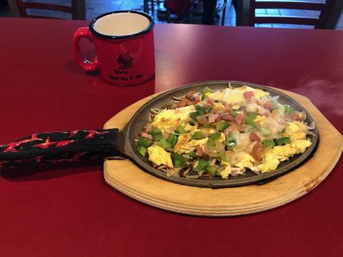 A sizzling skillet of scrambled eggs topped with green peppers, ham, and cheese, alongside a red mug on a table.