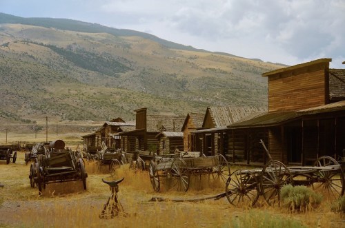 Abandoned wooden buildings and old wagons in a ghost town, set against a mountainous landscape under a cloudy sky.