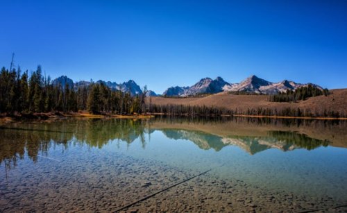 A serene lake reflects mountains and a clear blue sky, surrounded by trees and rugged terrain.