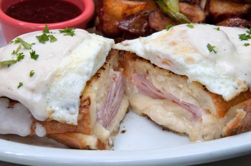 A close-up of a sandwich with ham and melted cheese, topped with a fried egg, served with roasted potatoes and dipping sauce.