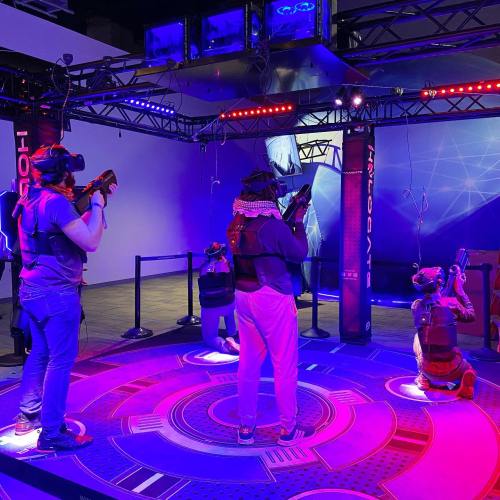 A group of people in VR headsets engaged in a virtual reality gaming experience in a colorful, high-tech environment.