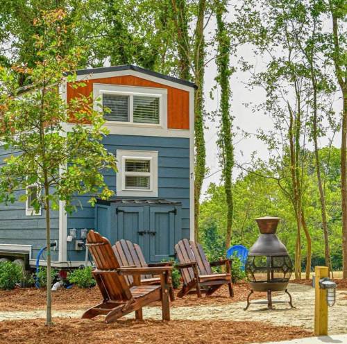 A cozy blue tiny house surrounded by trees, with wooden chairs and a fire pit in a serene outdoor setting.