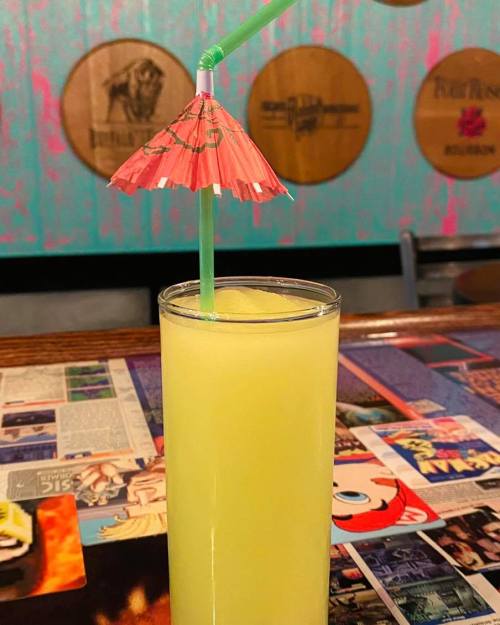 A tall glass of yellow drink with a pink umbrella straw, set against a colorful bar background.