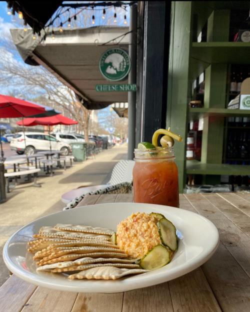 A plate of cheese spread with crackers and pickles, alongside a drink, viewed from a cozy café setting.