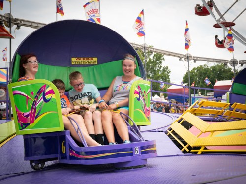 A group of four children and adults enjoy a ride at a colorful fair, surrounded by vibrant attractions and flags.