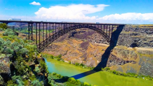 A large metal bridge arches over a river, surrounded by rocky cliffs and green vegetation under a blue sky.