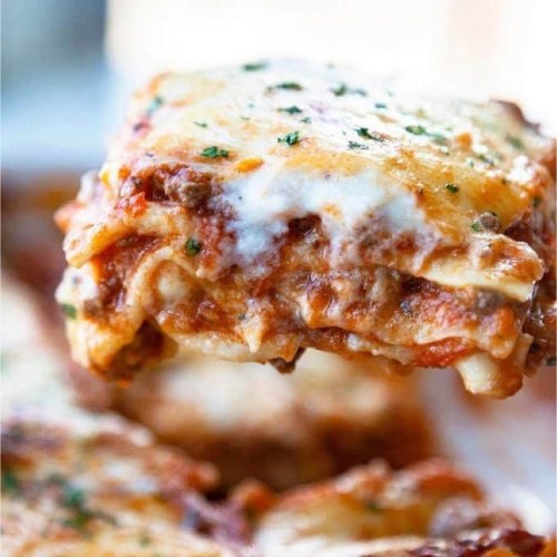 A slice of cheesy lasagna topped with herbs, held above a plate of more lasagna.
