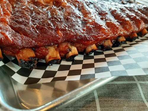 Close-up of a glazed barbecue rib rack on a checkered paper, resting on a metal tray.
