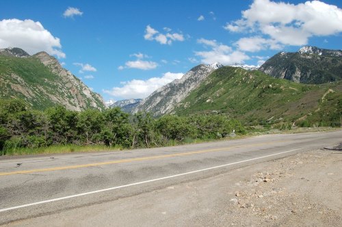 A scenic view of mountains and greenery alongside a road under a blue sky with fluffy clouds.