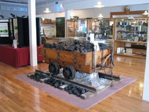 A wooden coal cart filled with coal, displayed on tracks in a museum exhibit with wooden floors and glass cases.