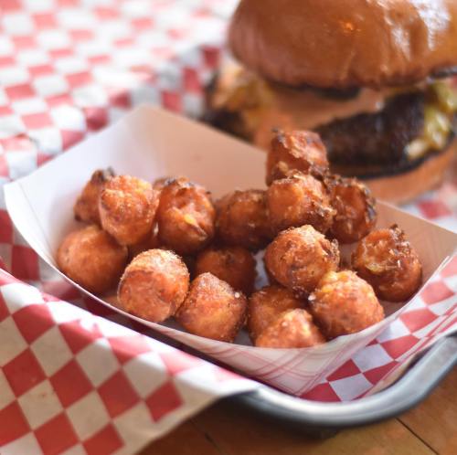 A serving of crispy tater tots in a paper tray, with a burger in the background on a checkered tablecloth.