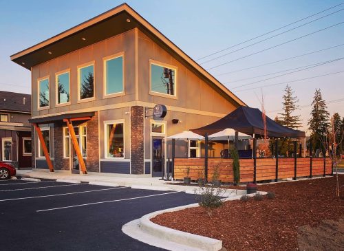 Modern restaurant exterior with large windows, outdoor seating, and a landscaped parking area at sunset.