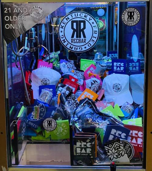 A claw machine filled with colorful merchandise, including hats and stickers, labeled "Kentucky Fried Rebar."