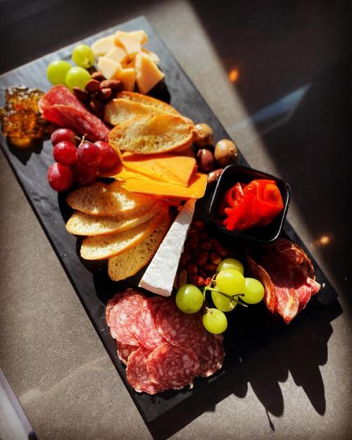 A charcuterie board featuring meats, cheeses, fruits, nuts, and a small bowl of red peppers on a dark slate platter.