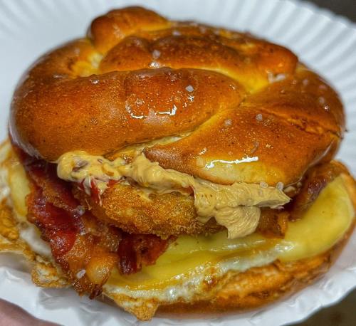 A close-up of a pretzel sandwich filled with bacon, cheese, and a creamy sauce, served on a paper plate.