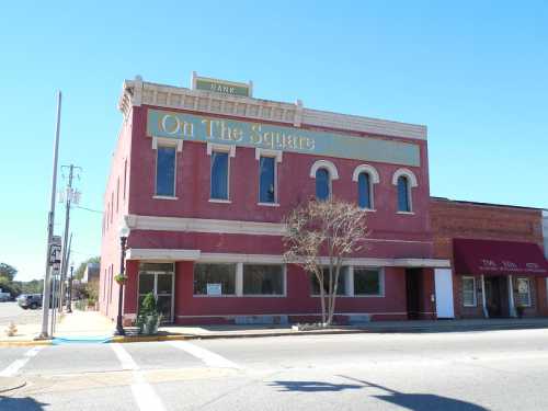 A red brick building with large windows, featuring the sign "On The Square" and a bank logo, on a sunny street.