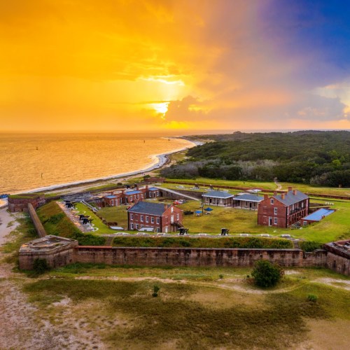 Aerial view of a historic fort by the coast at sunset, with buildings and lush greenery surrounding the area.