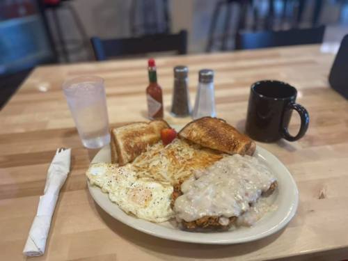 A hearty breakfast plate with eggs, hash browns, toast, chicken fried steak, gravy, and a coffee mug on a wooden table.