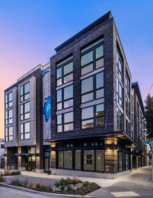 Modern multi-story building with large windows, featuring a mural and landscaped entrance at dusk.