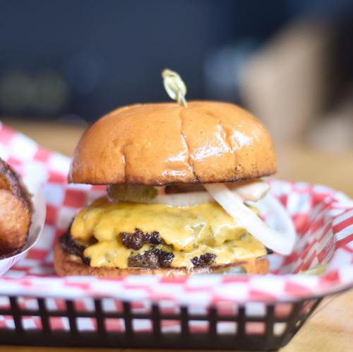 A close-up of a cheeseburger with two patties, melted cheese, pickles, and onions in a toasted bun, served in a basket.