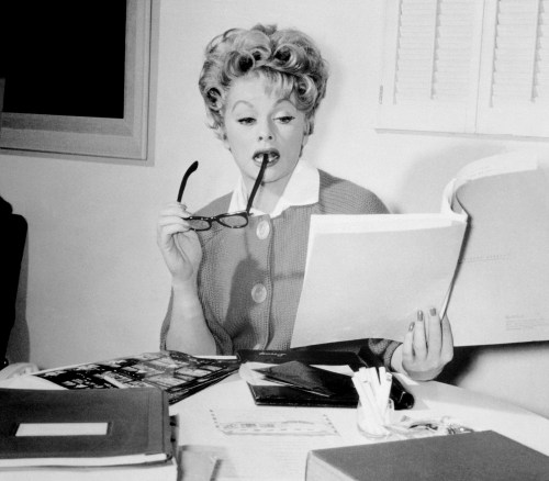 A woman in a vintage outfit reads documents while holding glasses in her mouth, surrounded by papers and folders.