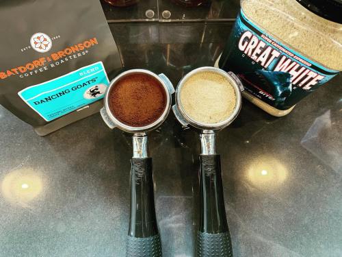 Two coffee portafilters filled with ground coffee, alongside bags of "Dancing Goats" coffee and "Great White" powder.