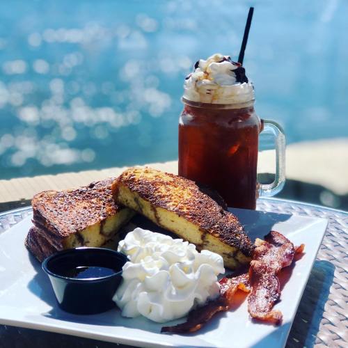 A plate of French toast with whipped cream, bacon, and syrup, alongside a jar of iced drink topped with whipped cream.