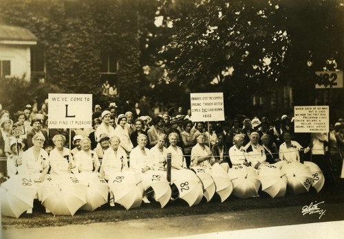A historic photo of women holding umbrellas and signs, gathered for a rally, promoting a cause in a public space.