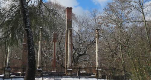 Snow-covered ruins with tall, weathered columns surrounded by bare trees and a blue sky.