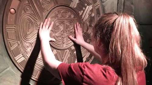 A person with long hair touches a large, intricate stone carving on a wall.
