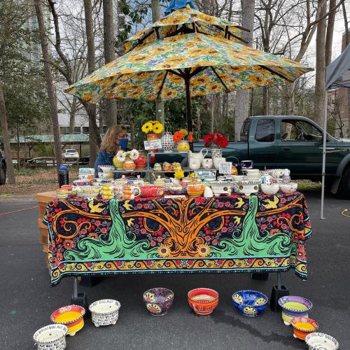 A colorful market stall with pottery and flowers, shaded by a vibrant umbrella, set in a wooded area.
