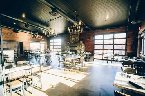 Bright, modern restaurant interior with wooden accents, large windows, and a stone fireplace. Tables are set for dining.
